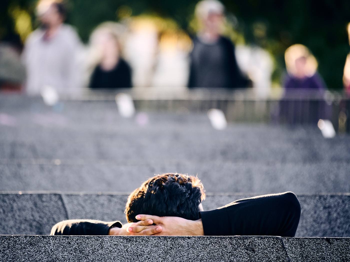 A person sitting on outdoor steps with hands behind their head, a blurred crowd walking past behind them